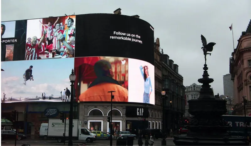 Curved LED screen across the upper floors of a corner building, above shops and a busy road, with street lamps in the foreground